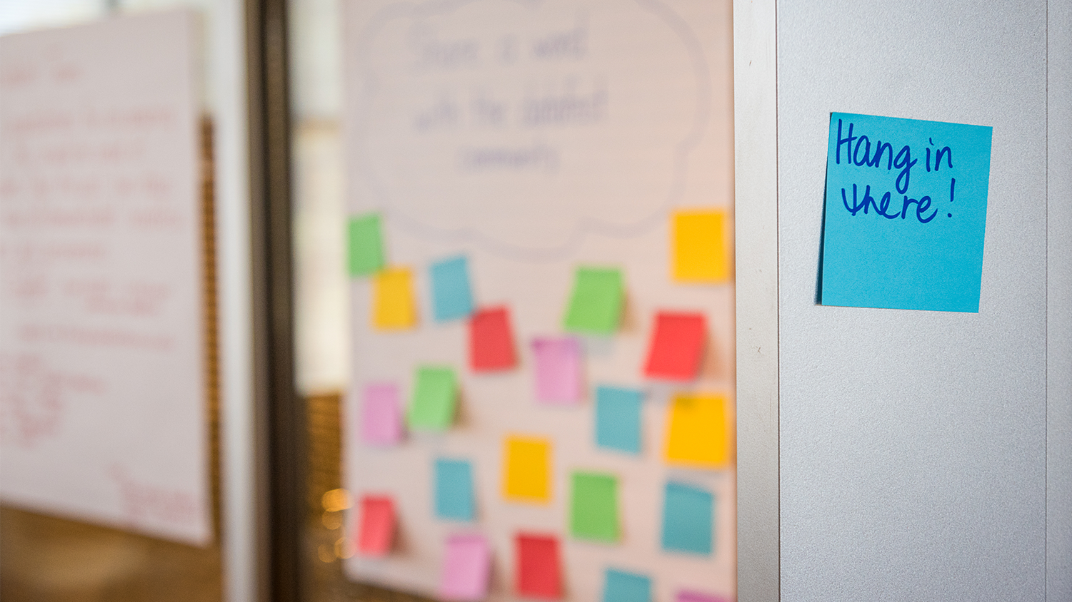 A blue sticky note with text "Hang in there" on a wall next to a posterboard with other sticky notes